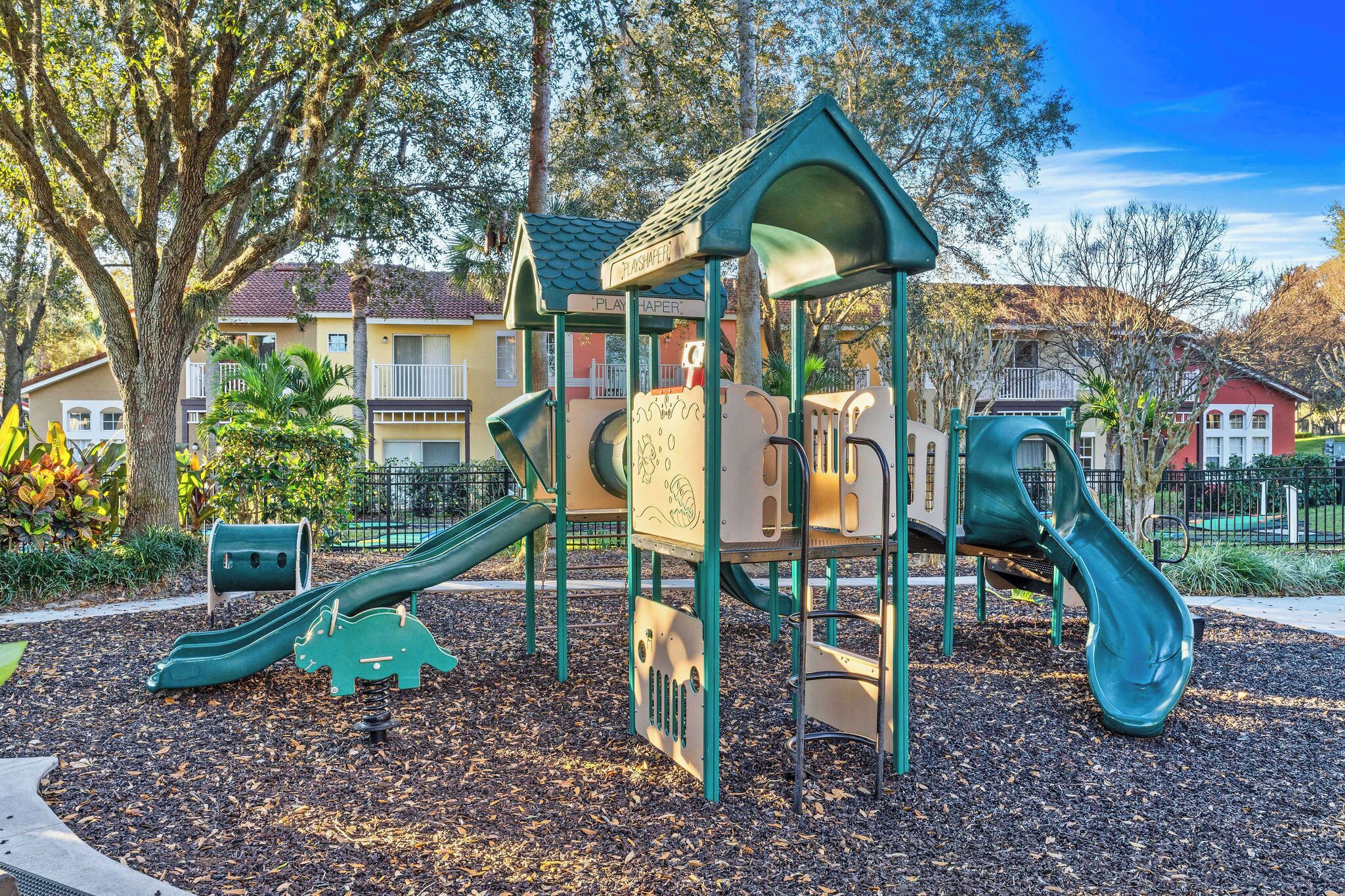 Ein Spielplatz mit Rutschen und Klettergerüsten, umgeben von Bäumen und Wohnhäusern.