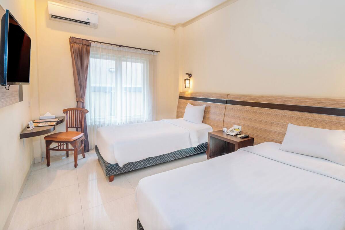 A hotel room with two beds, a desk, a chair, an air conditioner, a TV, and a window with curtains.