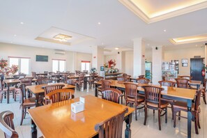 A spacious dining area with multiple wooden tables and chairs, a bar area, and a large window providing natural light. - Hotel Puri Indah Subak (Mataram)