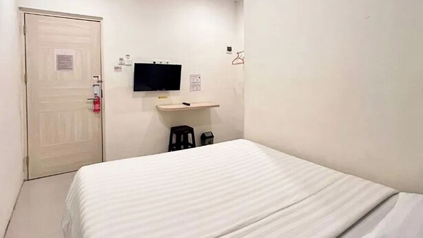 Een hotelkamer met een bed, een aan de muur bevestigde televisie, een klein krukje en een deur met een brandmelder.