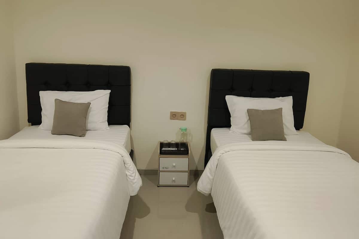 A hotel room with two beds, each with a black headboard, white bedding, and a single pillow. There is a nightstand with a bottle and a small drawer between the beds.