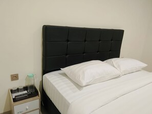 Una cama con cabecera negra, ropa de cama blanca y una mesita de noche con un control remoto y un recipiente de vidrio.