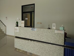 Un mostrador de recepción con encimera moteada, una planta en maceta, un espejo, un código QR y un letrero que indica la zona de registro.