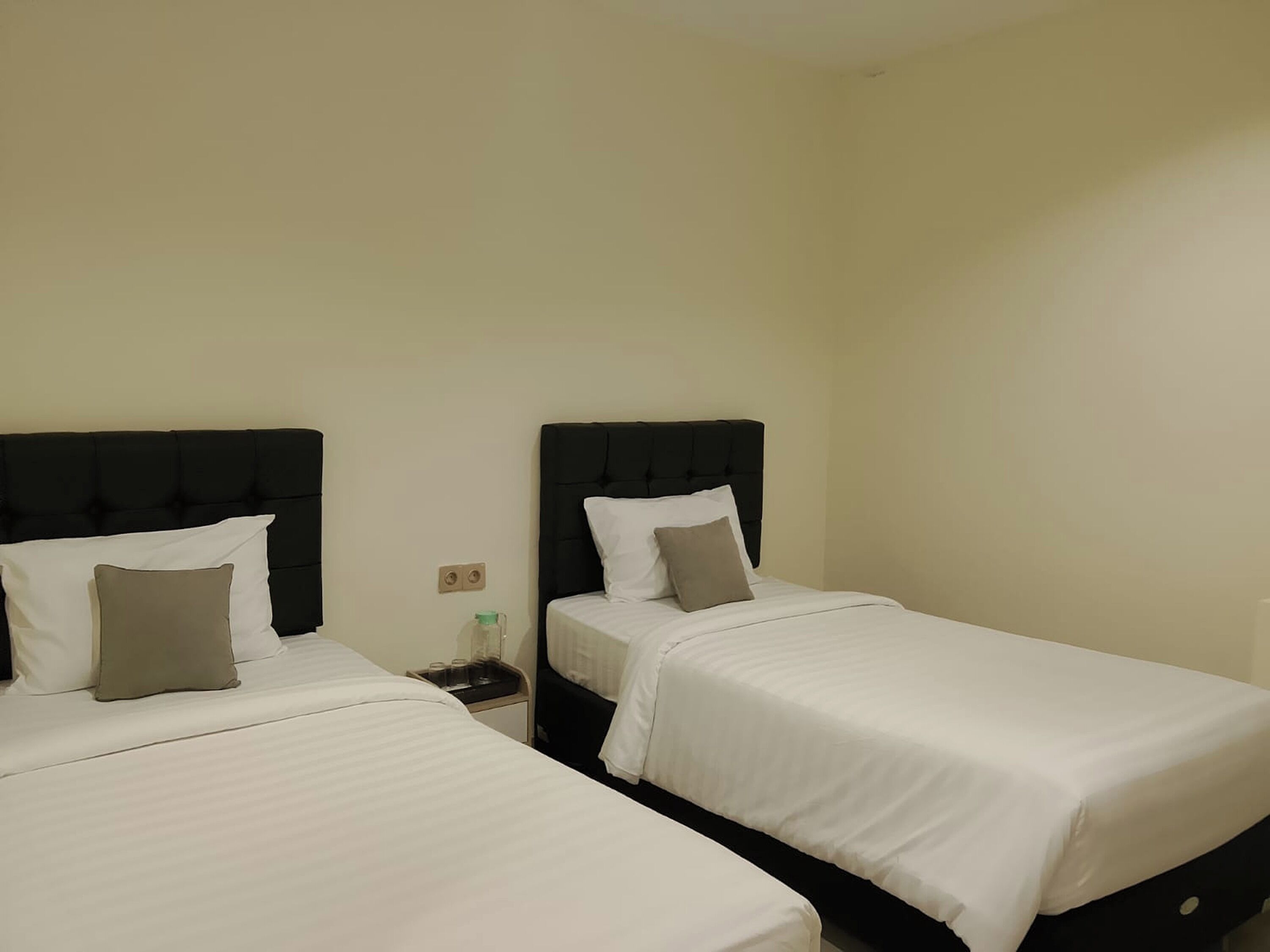A room with two beds, each with a black headboard and white bedding. There is a nightstand with a lamp and a bottle on it.