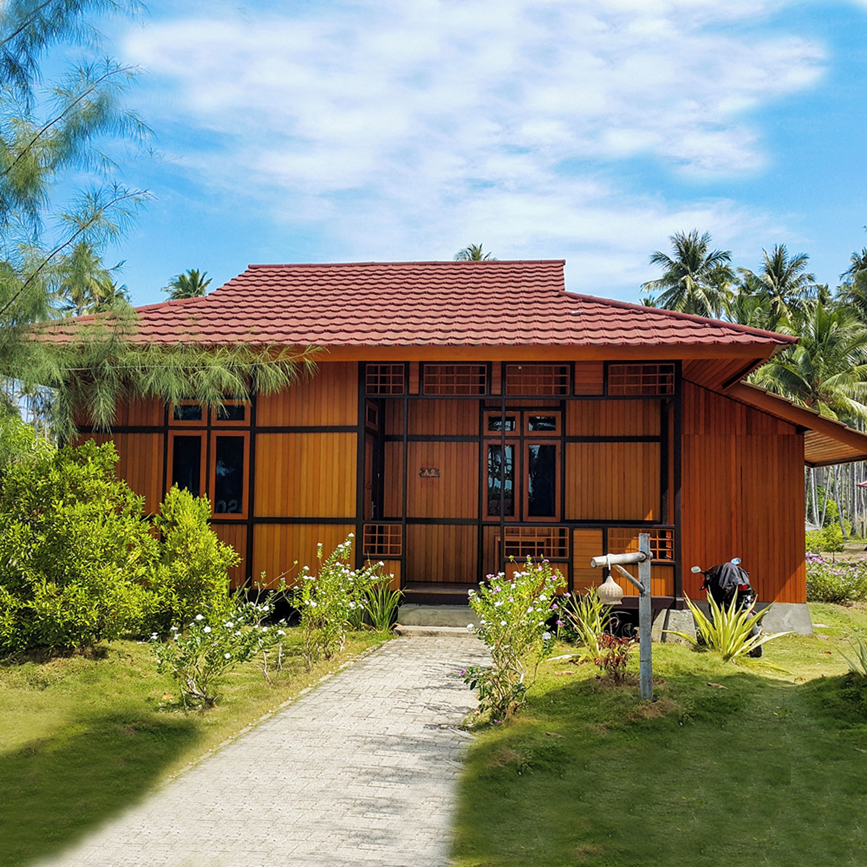 A wooden house with a tiled roof, surrounded by greenery and palm trees.