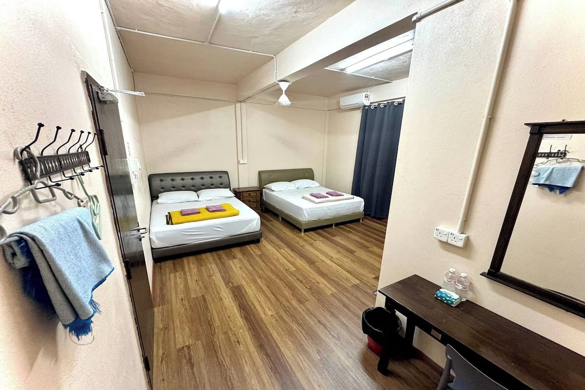 A hotel room with two beds, a wooden floor, a desk with a mirror, and a wall-mounted air conditioning unit.