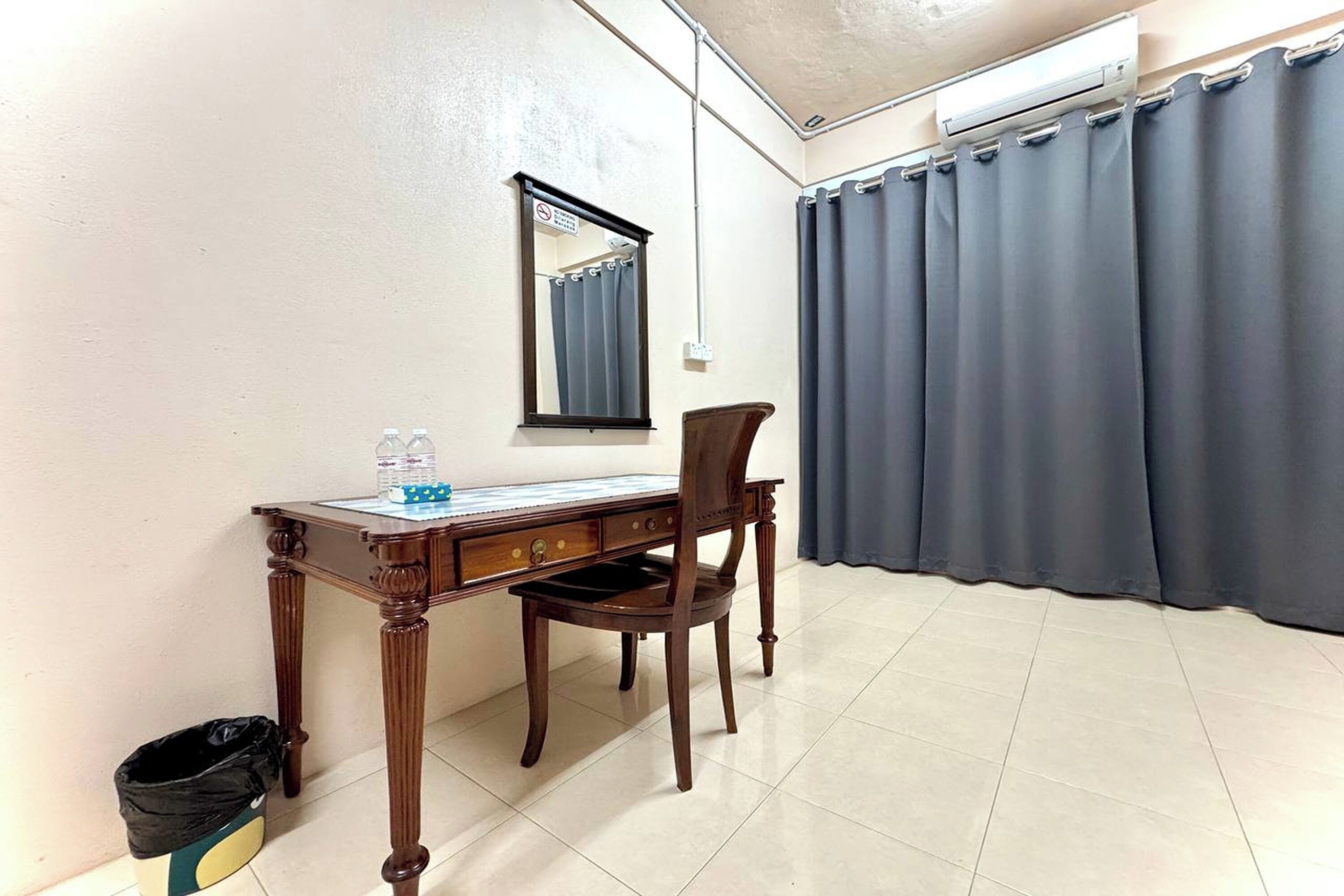 Una habitación con un escritorio de madera, una silla, un espejo, un ventanal con cortinas y un cesto de basura.