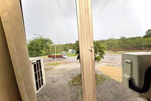 A view through a window with a metal frame, a gravel area, a road with vehicles, and greenery in the background.
