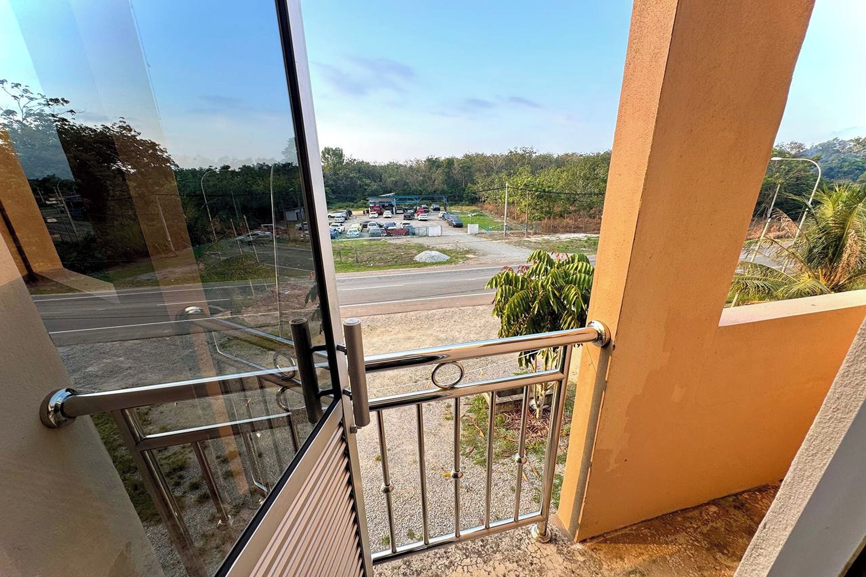 Vista desde un balcón con baranda de metal, un estacionamiento y follaje al fondo.