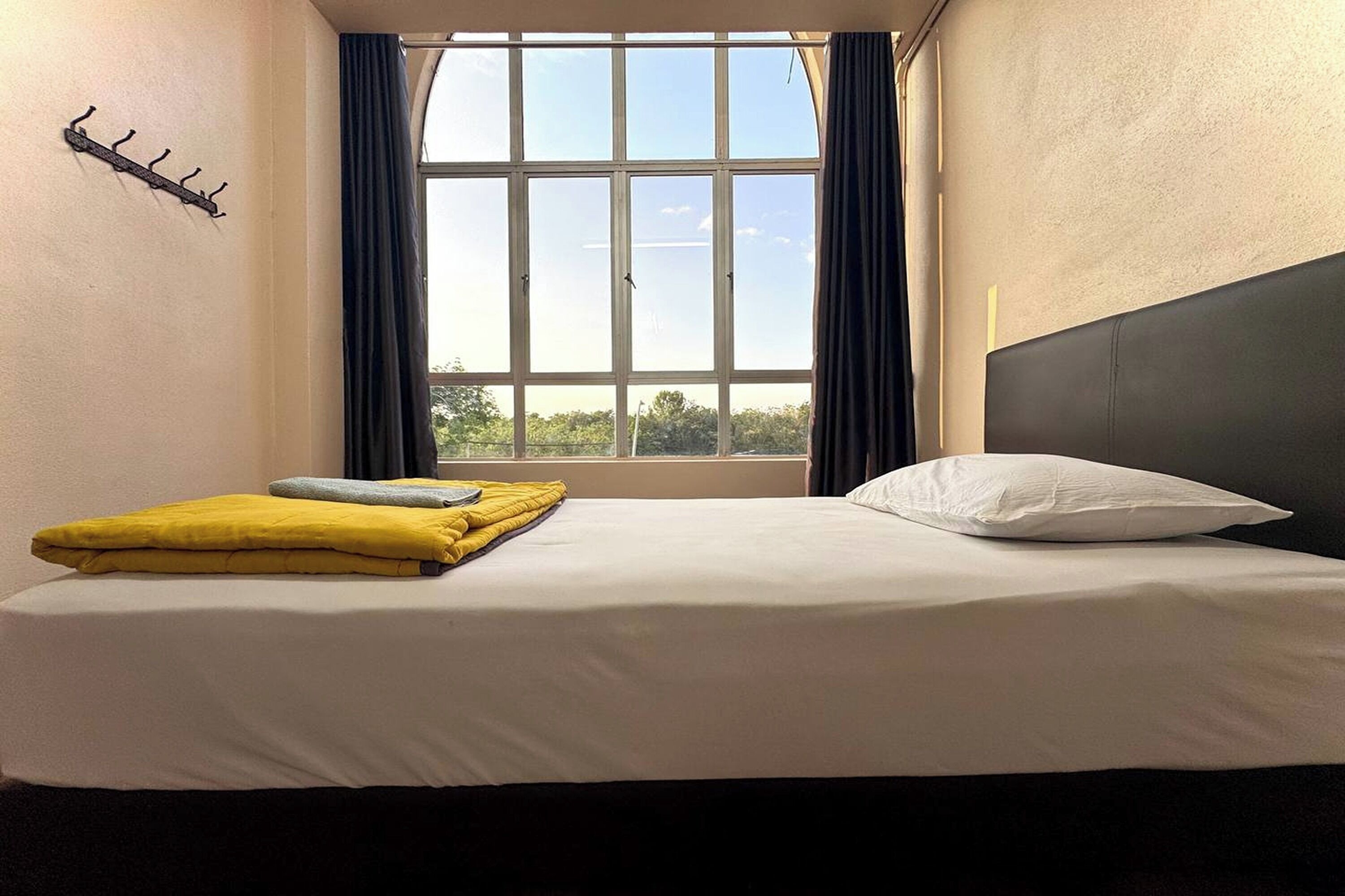 Habitación de hotel con cama, ventana con cortinas, perchero y vista a zonas verdes.
