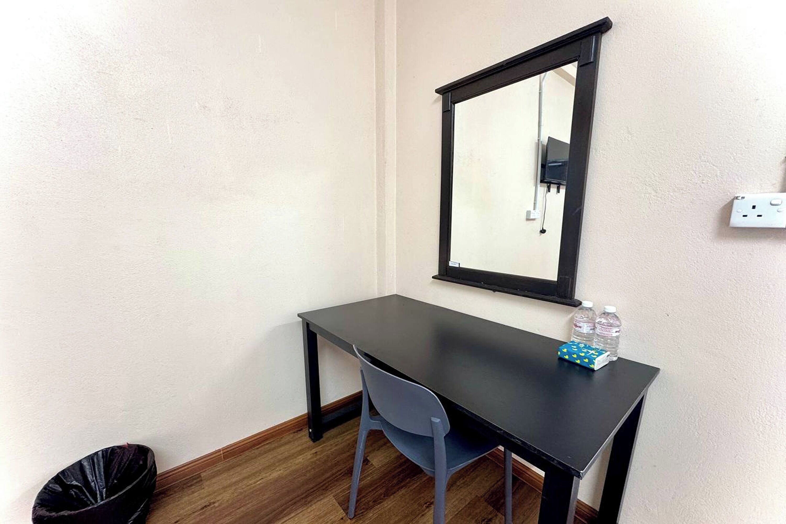 Una habitación pequeña con un escritorio, una silla y un espejo. Hay un cubo de basura y una botella de agua sobre el escritorio.