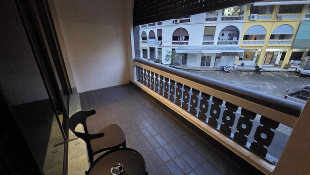 A balcony with a chair and a table, a railing, and a view of a building with multiple stories and balconies. - Terminal Garage (Phuket)