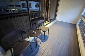 A balcony with two chairs and a small table, a railing, and a view of a building with arches. - Terminal Garage (Phuket)