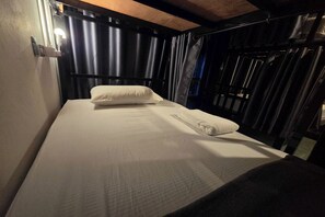A single bed with a white mattress and pillow, a small bedside table, and a window with curtains. - Terminal Garage (Phuket)