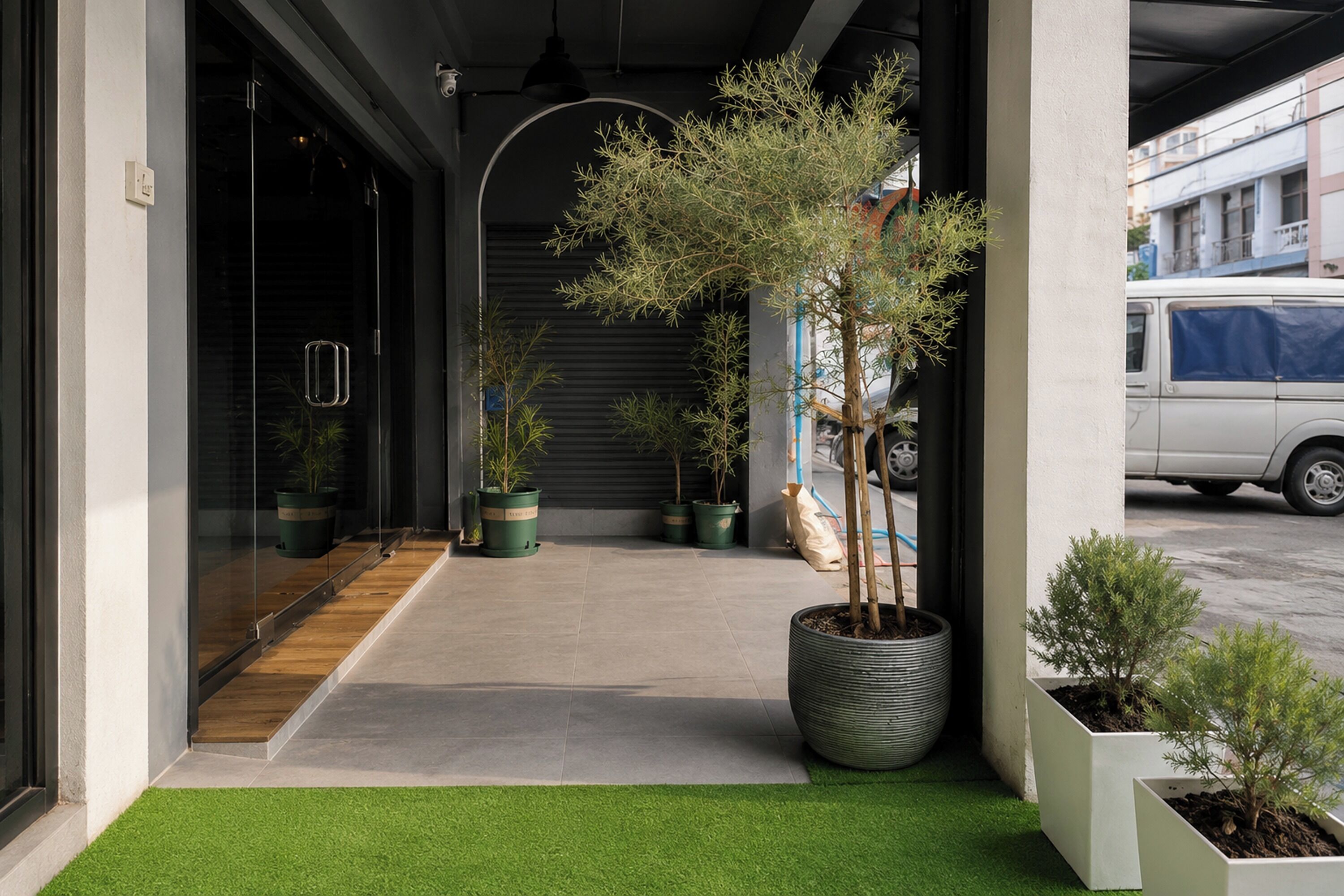 Una entrada de edificio moderna con césped artificial, plantas en macetas y un vehículo estacionado afuera.