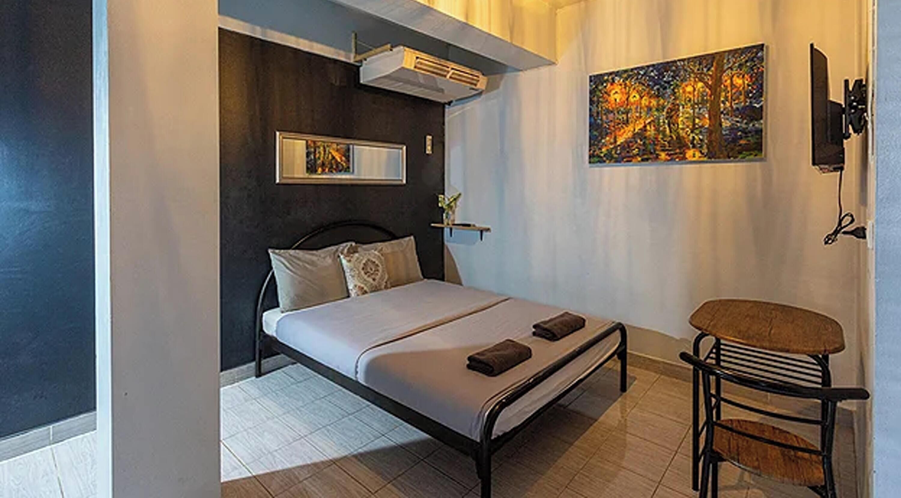 A bedroom with a bed, bedside table, chair, artwork, and an air conditioning unit.