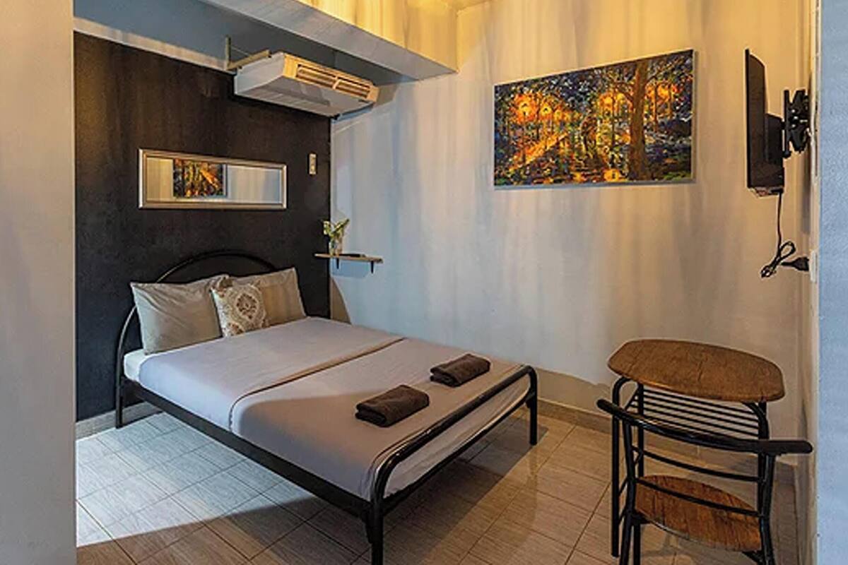 Un dormitorio con cama, mesita de noche, silla, una obra de arte y un equipo de aire acondicionado.