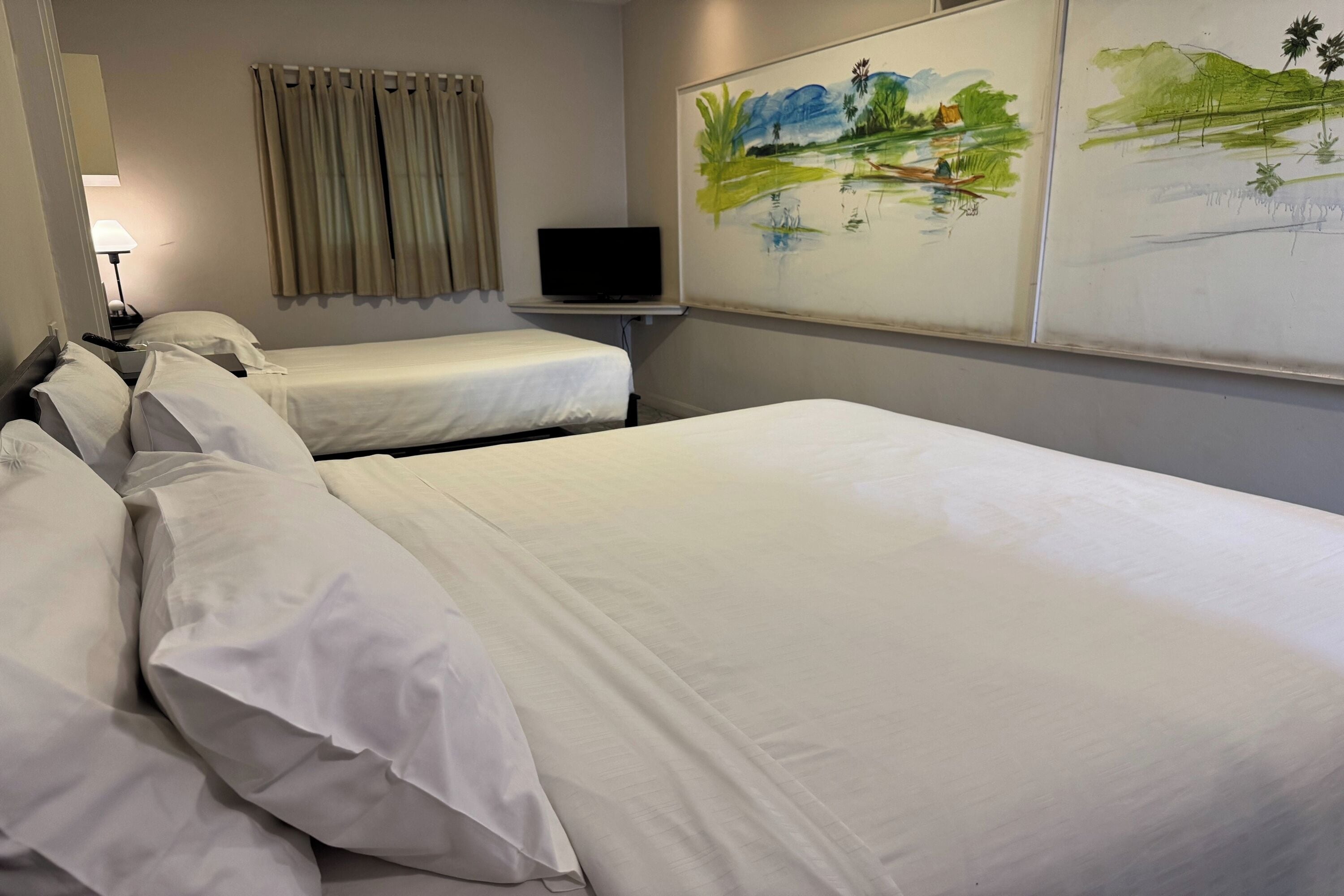 A hotel room with two beds, a television on a stand, a painting on the wall, and a window with curtains.
