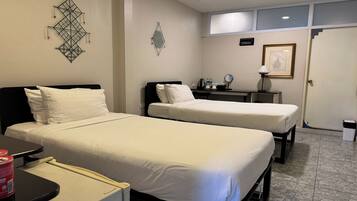 A hotel room with two beds, a desk, a chair, a small table, a lamp, and a framed picture on the wall.