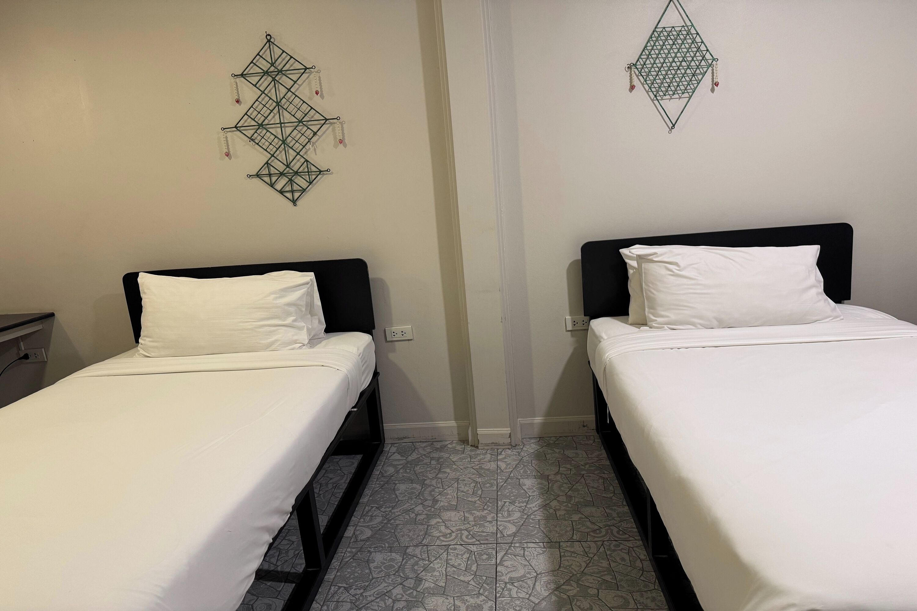 Two single beds with black headboards and white bedding, a wall-mounted geometric decoration, and patterned floor tiles.