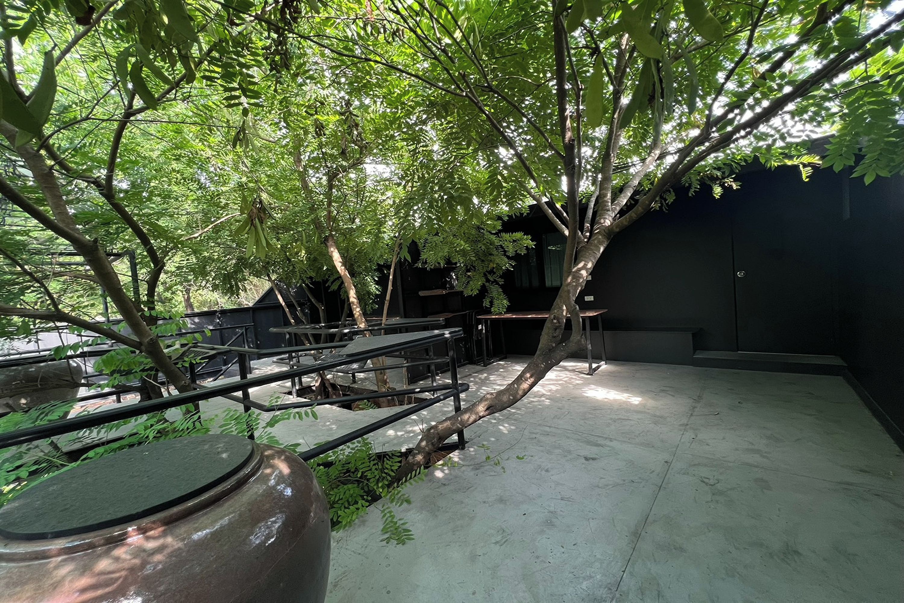 An outdoor area with a concrete floor, metal railings, and a variety of trees providing shade.