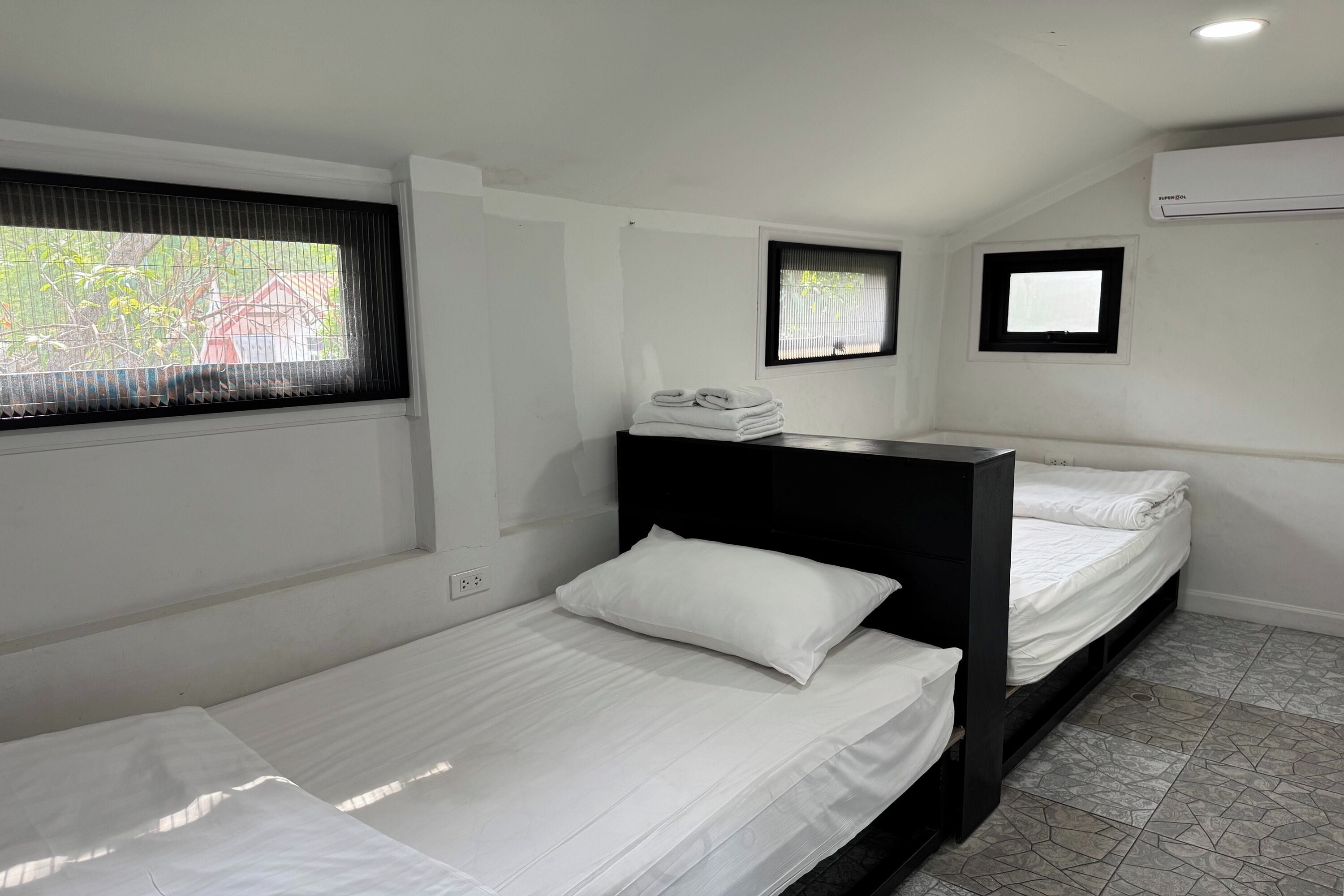 A room with two beds, a window with blinds, an air conditioning unit, and a small window with a view.