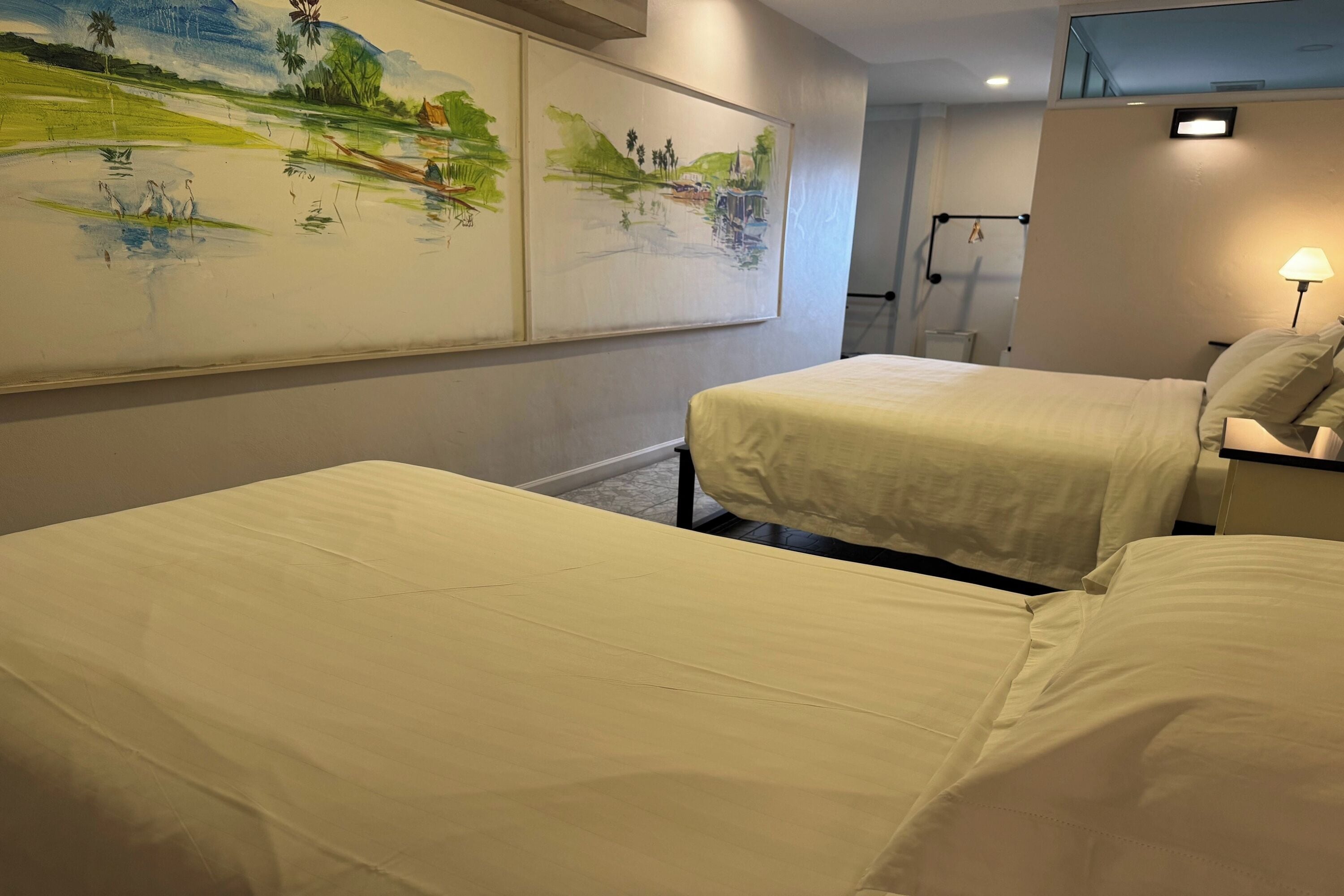 A hotel room with two beds, a painting on the wall, a lamp, and a small table.