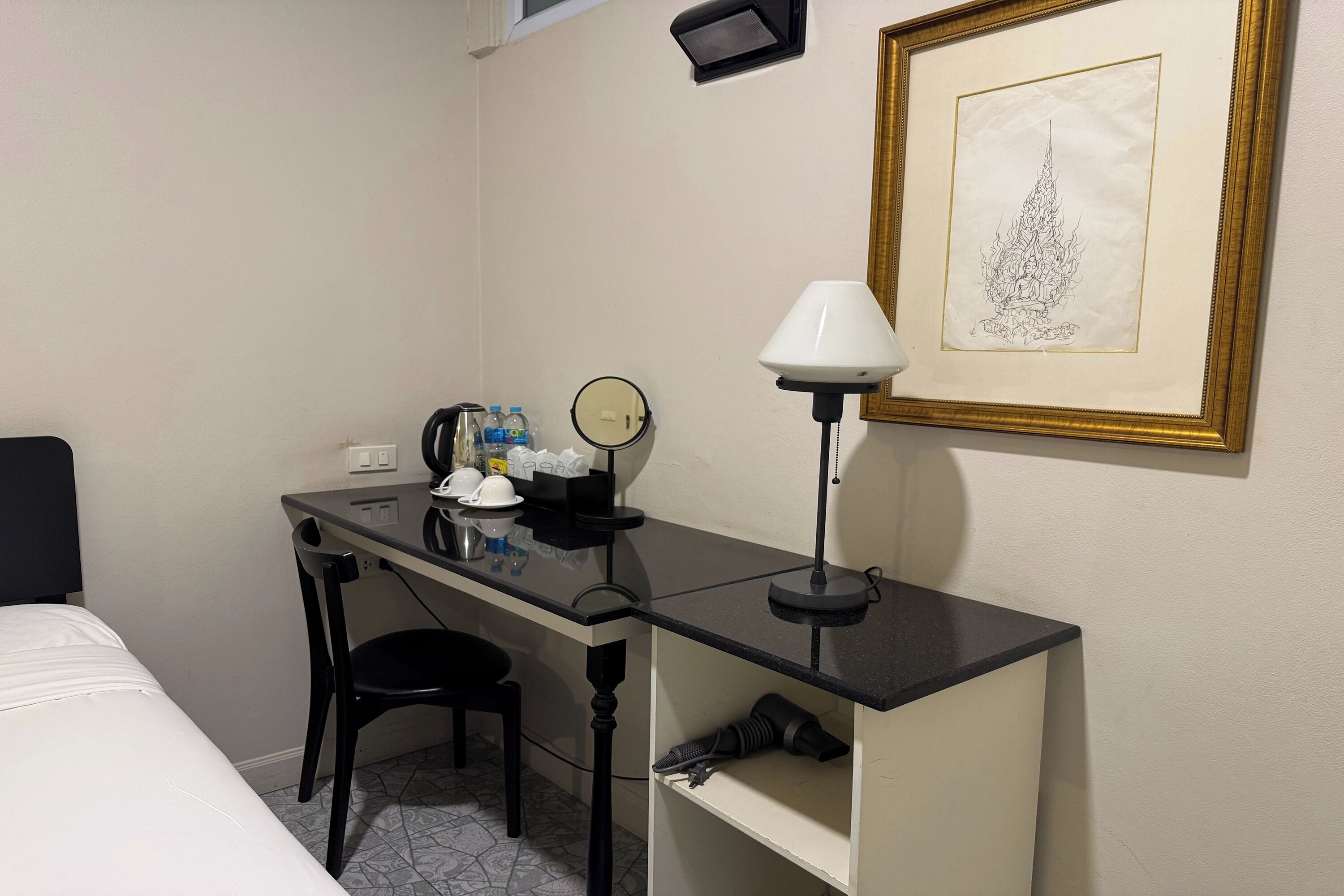 A hotel room with a bed, a desk with a chair, a lamp, a framed picture, and a shelf with a camera.