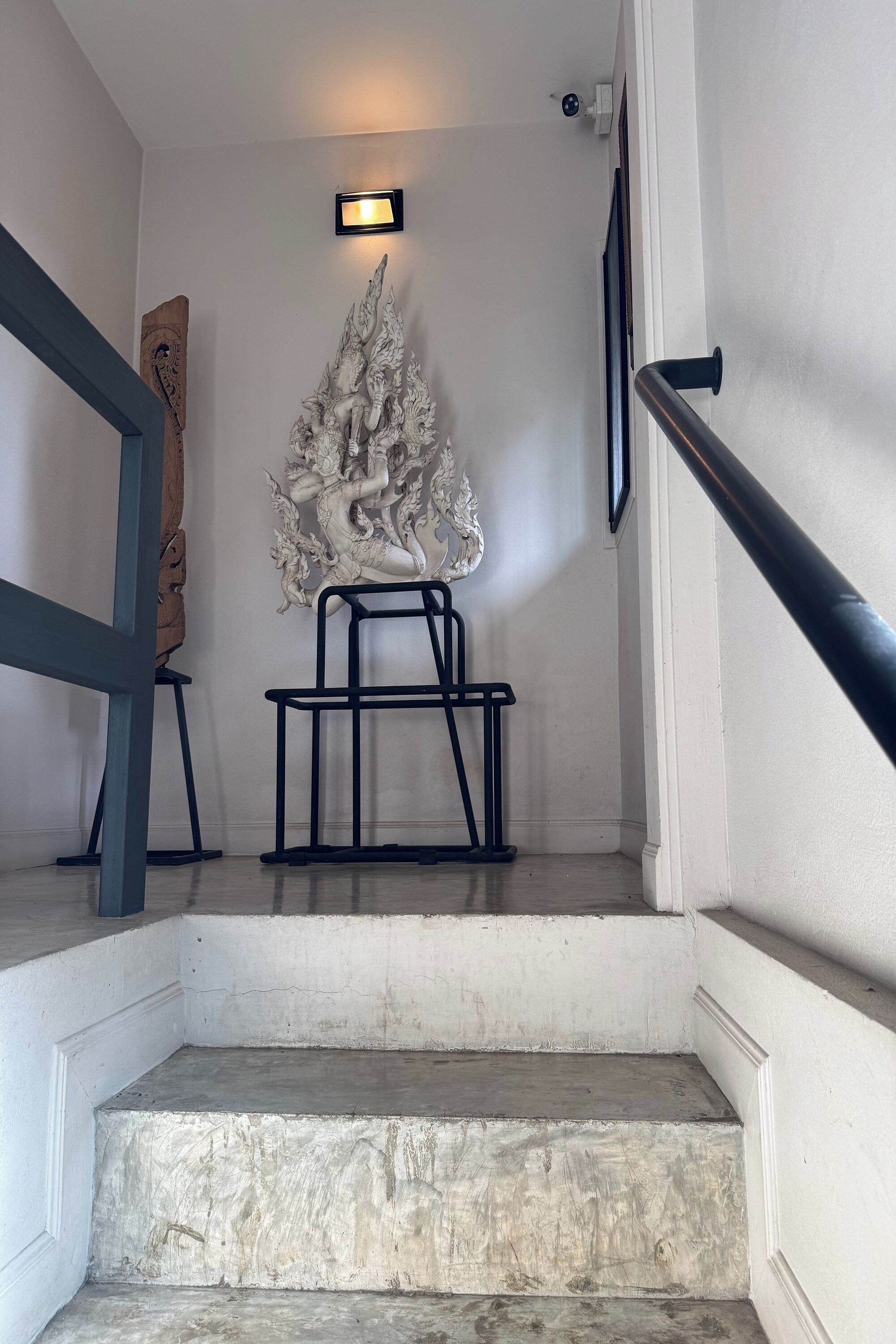 A staircase with a white wall, a black metal chair, a decorative white sculpture, and a small wall-mounted light fixture.
