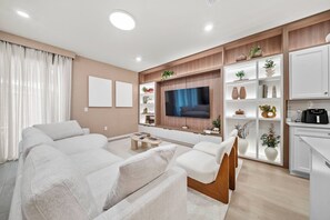 Un soggiorno moderno con una grande TV a schermo piatto, un divano, un tavolino da caffè e scaffali a muro con oggetti decorativi.