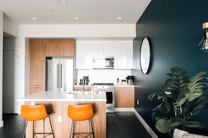 A modern kitchen with wooden cabinets, stainless steel appliances, and a central island with two orange bar stools. - Top-Floor 1BR Loft | Skyline Views | Walkable LoHi Denver (Denver)