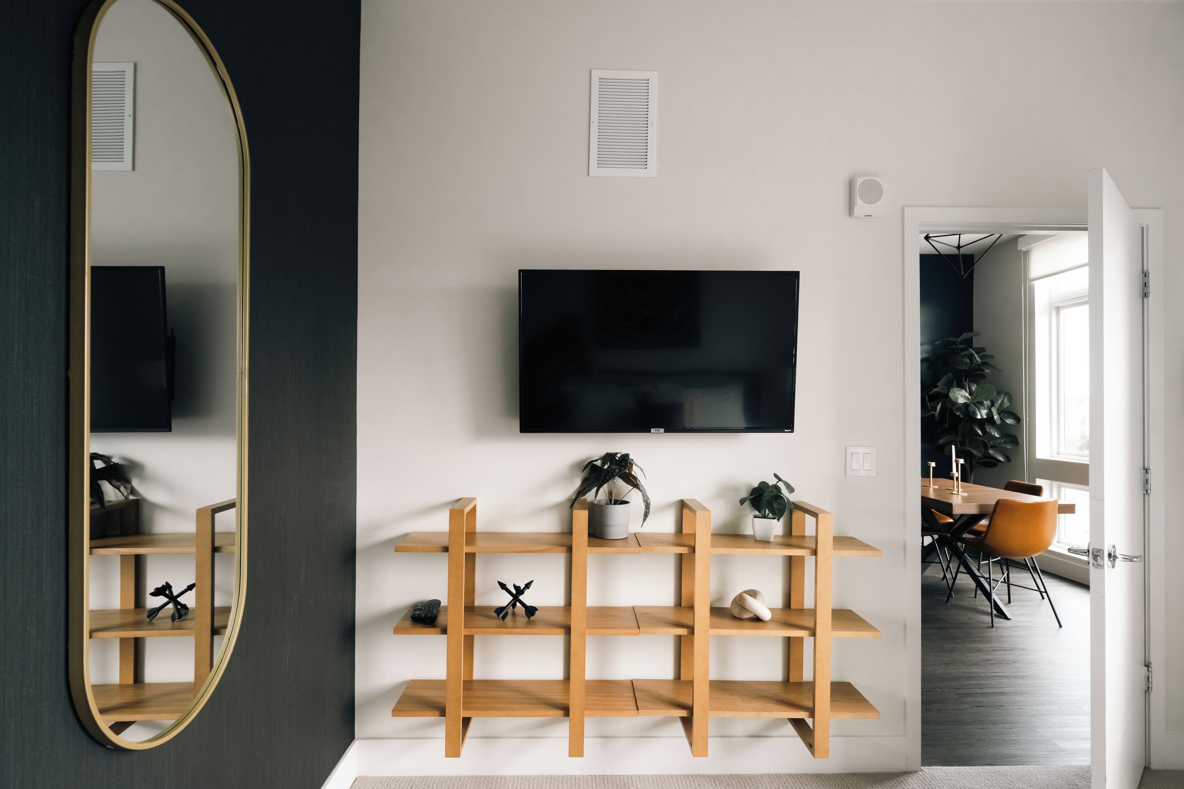 Una sala de estar moderna con un espejo grande, estanterías de madera, un televisor de pantalla plana y un comedor con mesa y sillas.