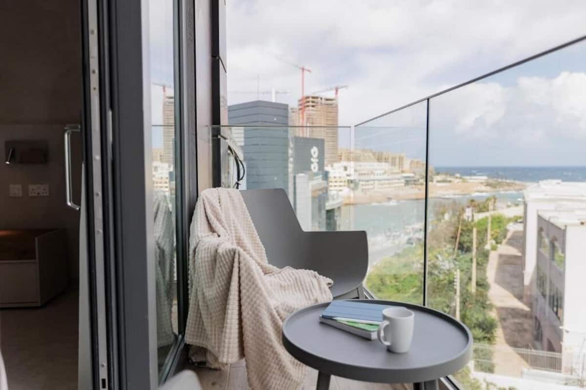 Un balcón con vistas a la ciudad y al océano, una silla con una manta, y una mesita con un libro y una taza.