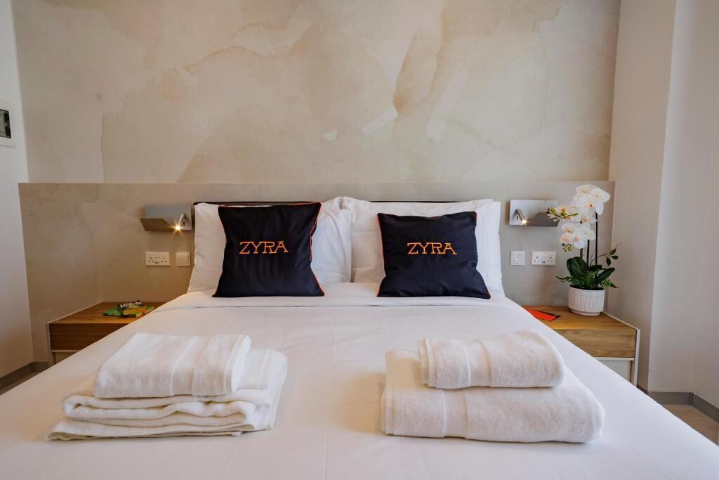 A hotel room with a bed, two pillows with 'ZYRA' text, a nightstand with a plant, and neatly folded towels.