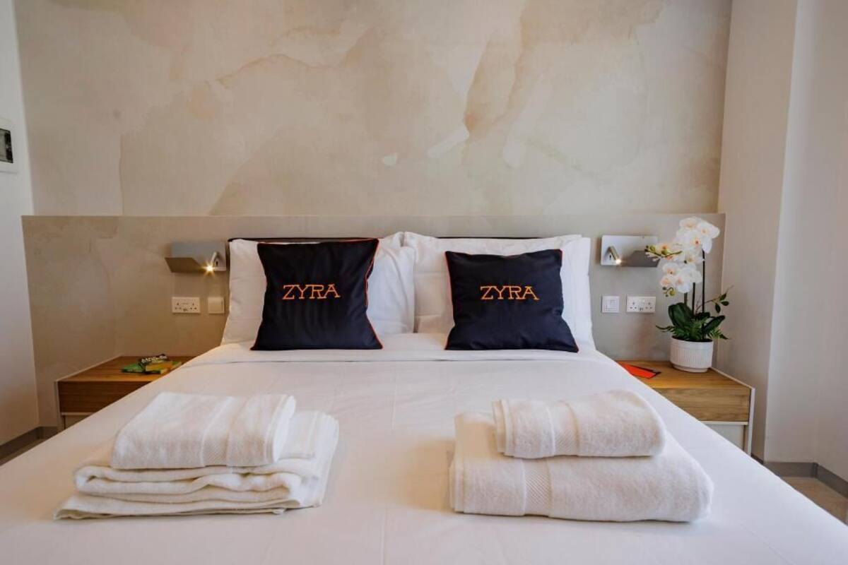 Habitación de hotel con cama, dos almohadas con el texto 'ZYRA', una mesita de noche con una planta y toallas dobladas con cuidado.