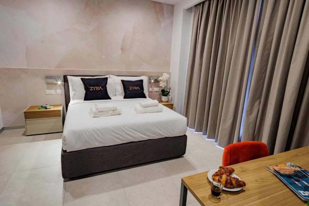 Una habitación de hotel moderna con una cama grande, una mesita de noche con lámpara, una pequeña mesa con desayuno y una ventana con cortinas.
