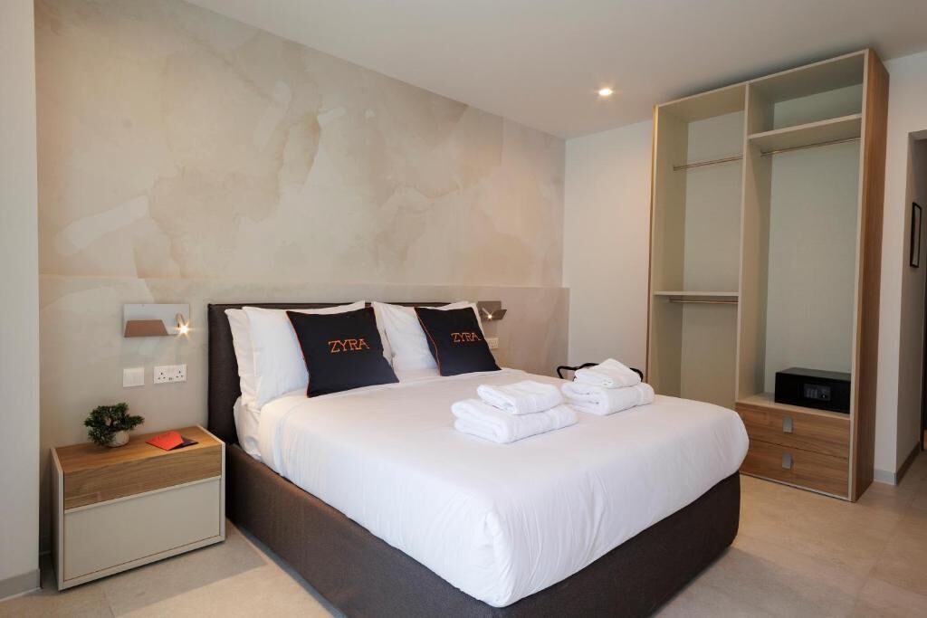 A modern hotel room with a large bed, a nightstand, a wardrobe, and a wall-mounted lamp.