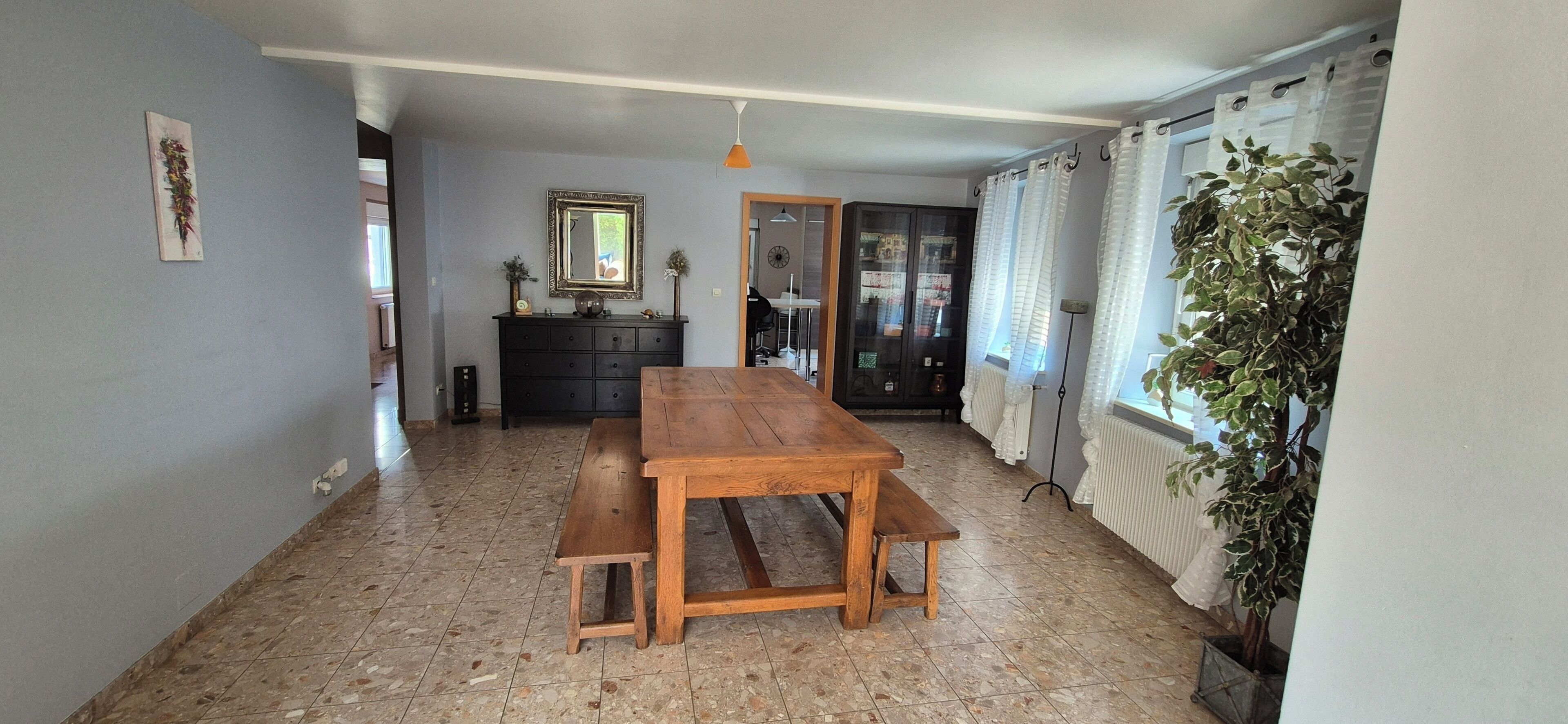 A dining area with a wooden table and benches, a dark-colored dresser, a mirror, and a potted plant.