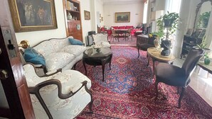 Ein Wohnzimmer mit einer Couch, Sesseln, einem Couchtisch, einem Esstisch mit Stühlen, einem Bücherregal und einem großen Fenster.