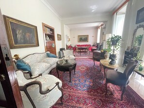 Ein Wohnzimmer mit einer Couch, Sesseln, einem Couchtisch, einem Esstisch mit Stühlen, einem Bücherregal und einem großen Fenster.
