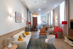 Ein modernes Hotelzimmer mit einer Couch, Sesseln, einem kleinen Tisch, einem Spielzelt und einem Fernseher.