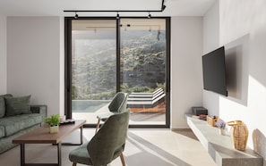 A modern living room with a large sliding glass door, a flat-screen TV mounted on the wall, a sofa, a chair, and a small table with a plant.