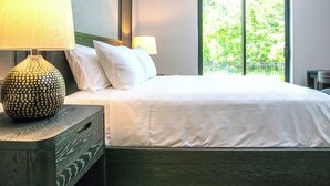 A neatly made bed with white linens, a wooden nightstand with a lamp, and a window with a view of greenery.