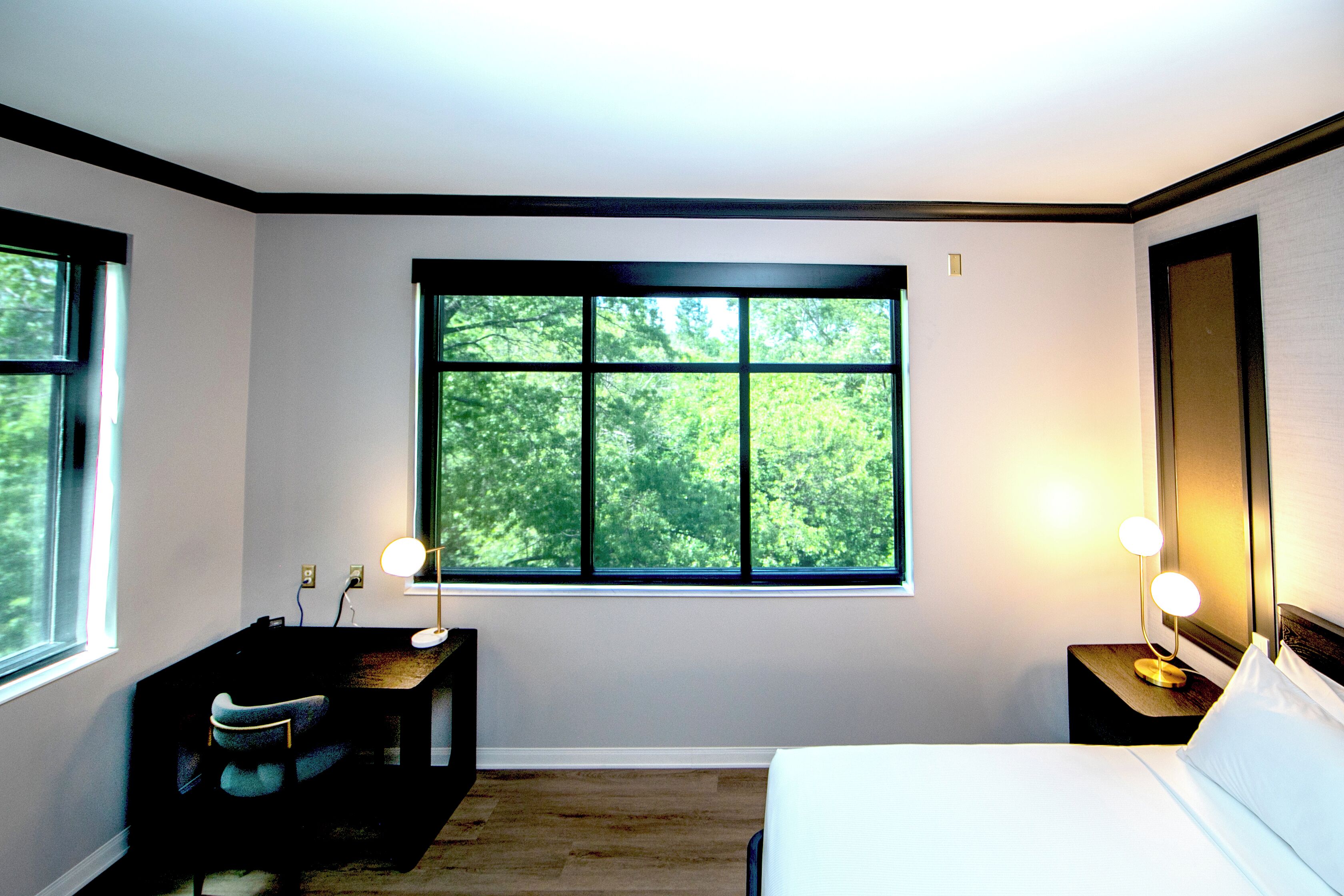A bedroom with a large window, a desk, a chair, a bedside table with lamps, and a bed.