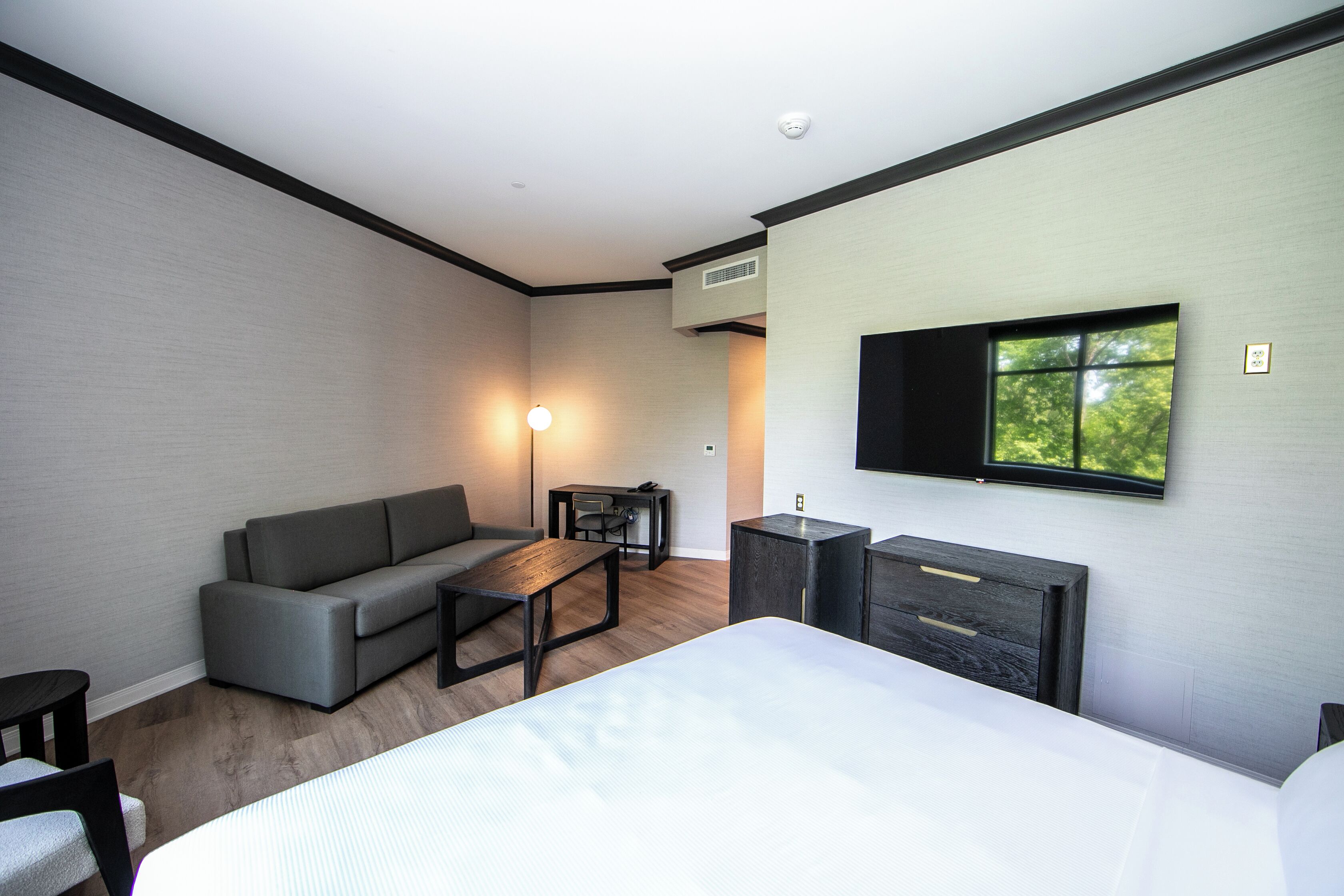 A modern hotel room with a bed, a sofa, a small table, a TV mounted on the wall, and a window.