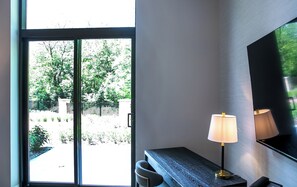 A room with a sliding glass door leading to a garden, a desk with a lamp, and a large mirror on the wall.