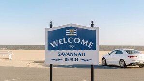 Un letrero de bienvenida a Savannah Beach, con cielo despejado y un coche estacionado al fondo.