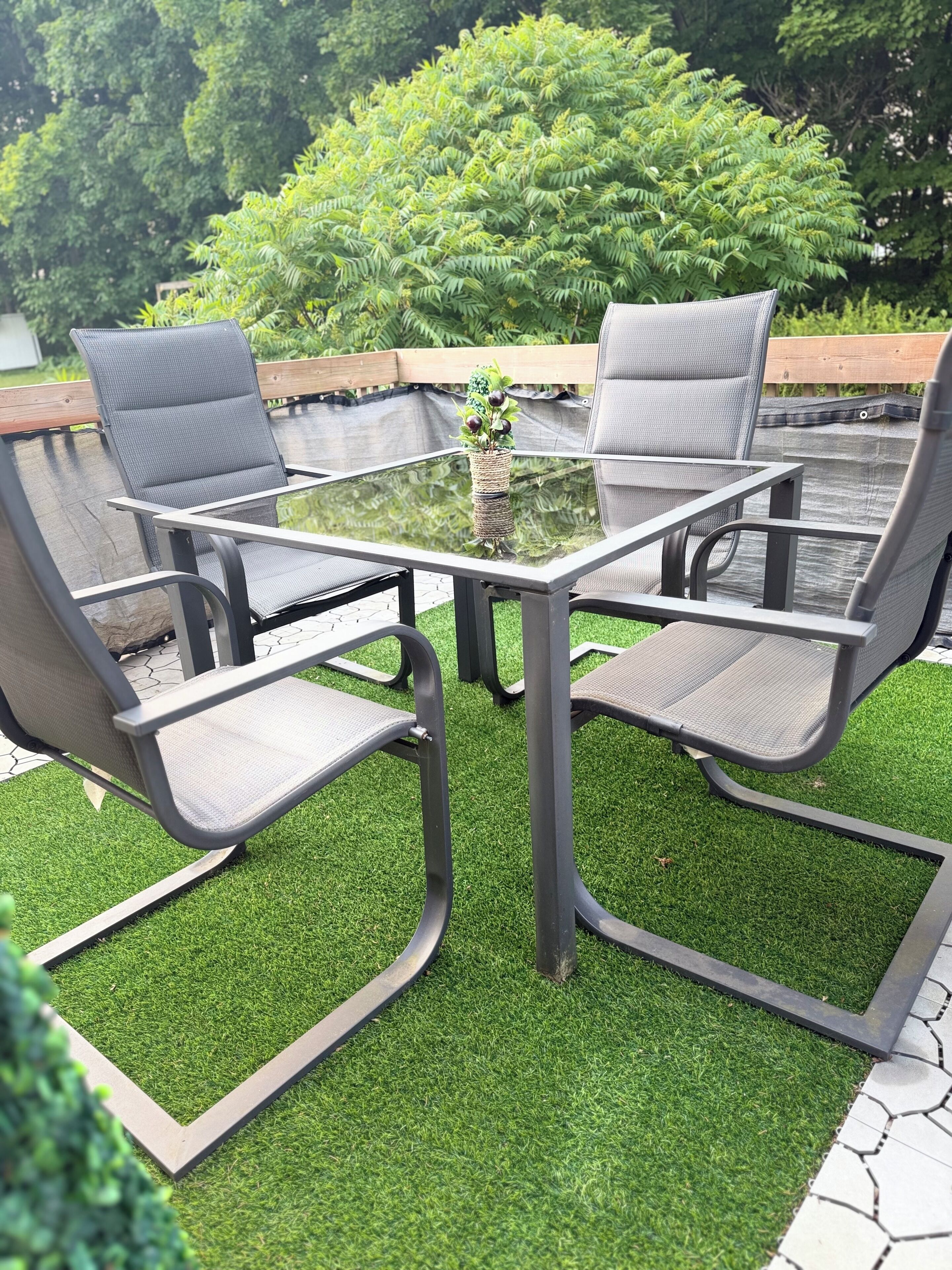 Une table à plateau en verre, accompagnée de quatre chaises, entourée de gazon synthétique, avec une terrasse en bois et un feuillage dense en arrière-plan.