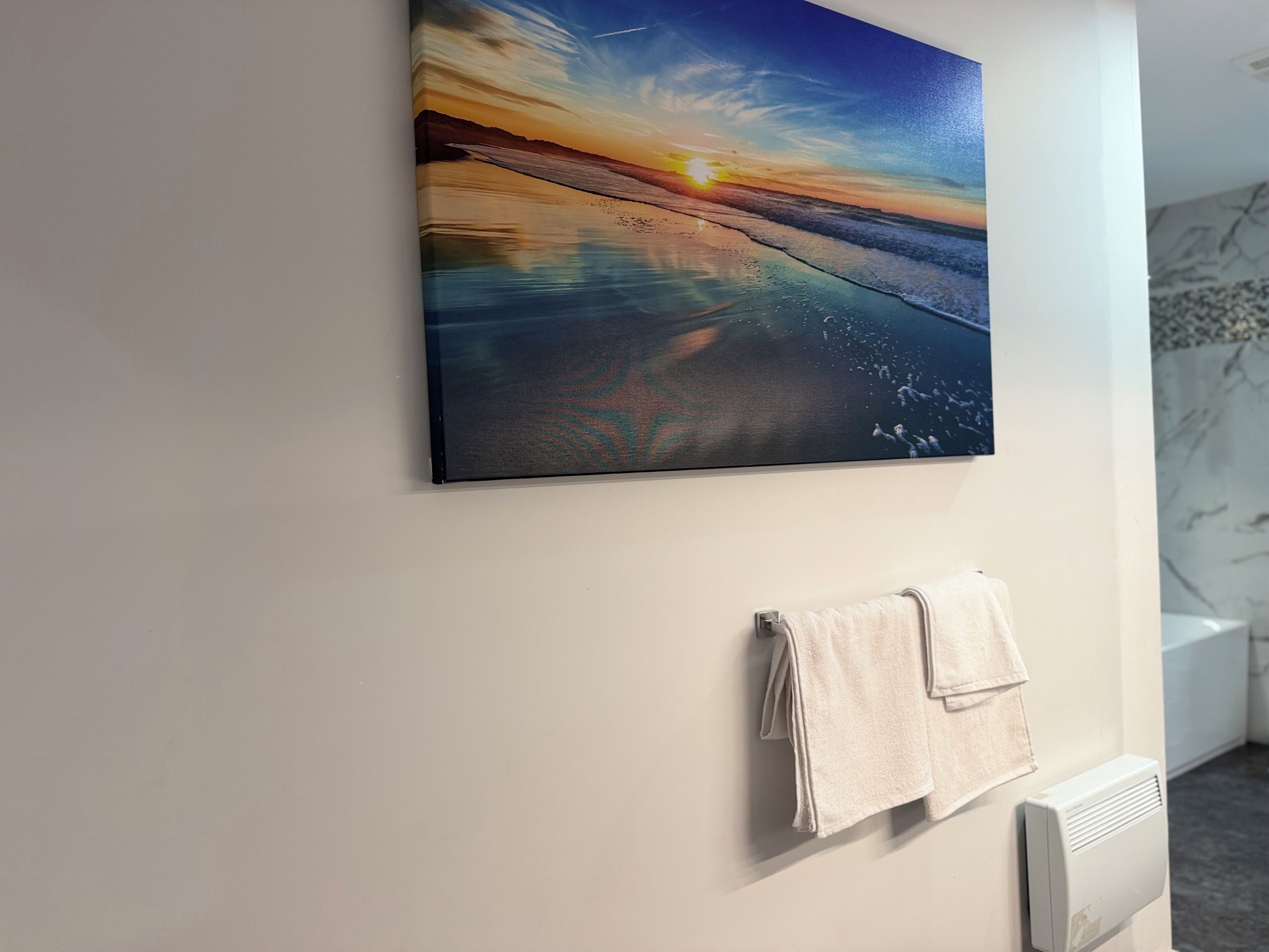 Un tableau représentant un coucher de soleil sur une plage, accroché au mur, une serviette posée sur un porte-serviettes et un radiateur.
