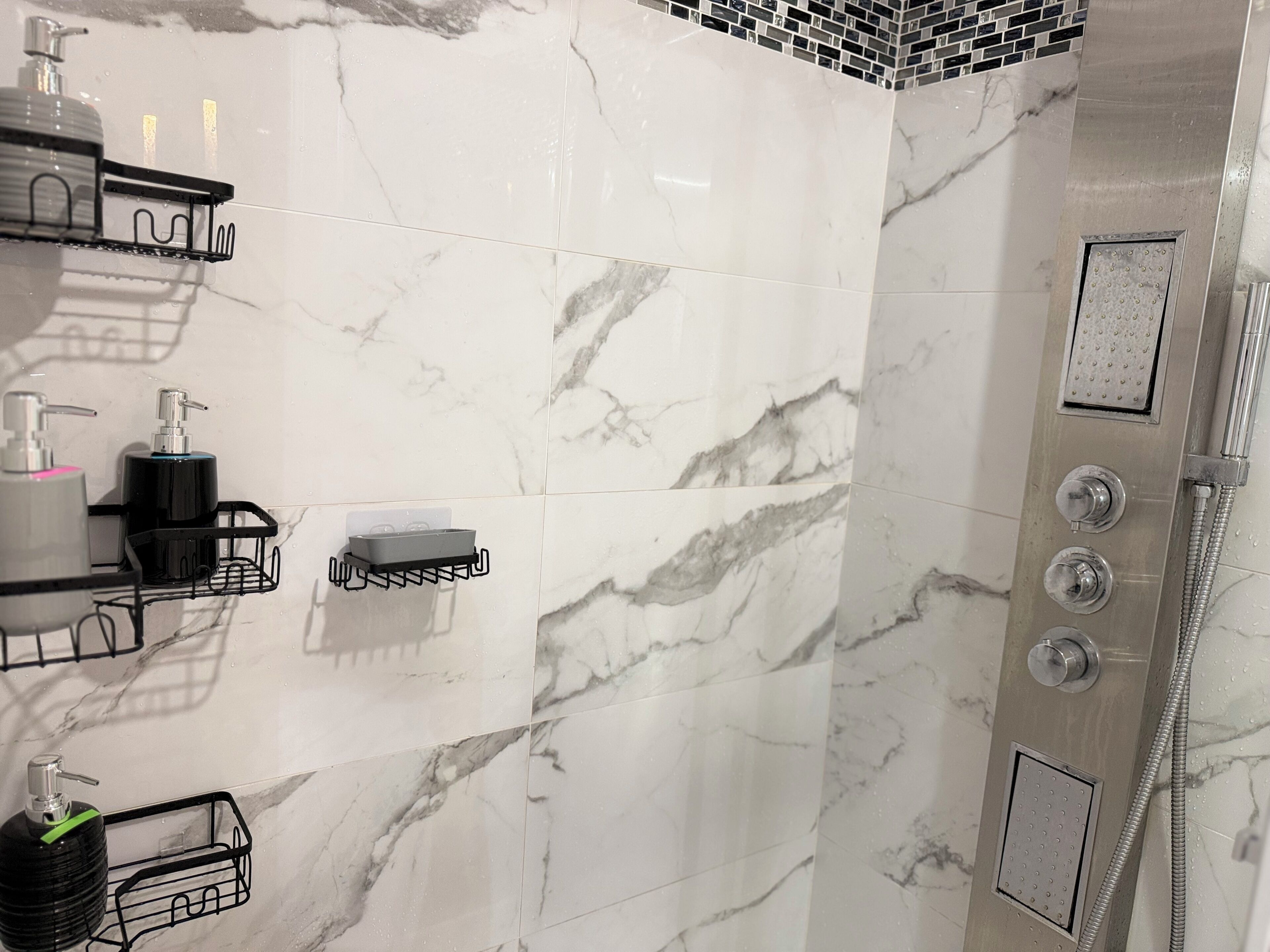 Une salle de bain aux murs recouverts de marbre, une douche dotée de plusieurs commandes et des étagères noires fixées au mur pour ranger les articles de toilette.